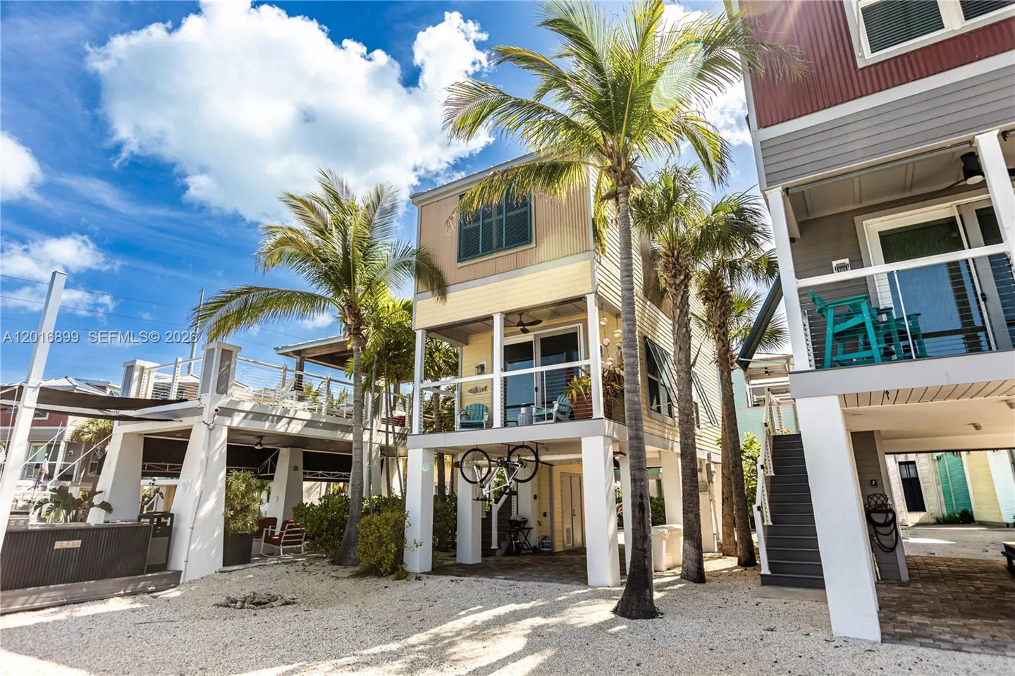 Exterior view of stilt houses with palm trees, under a blue sky with white clouds. A bicycle hangs under the yellow house.