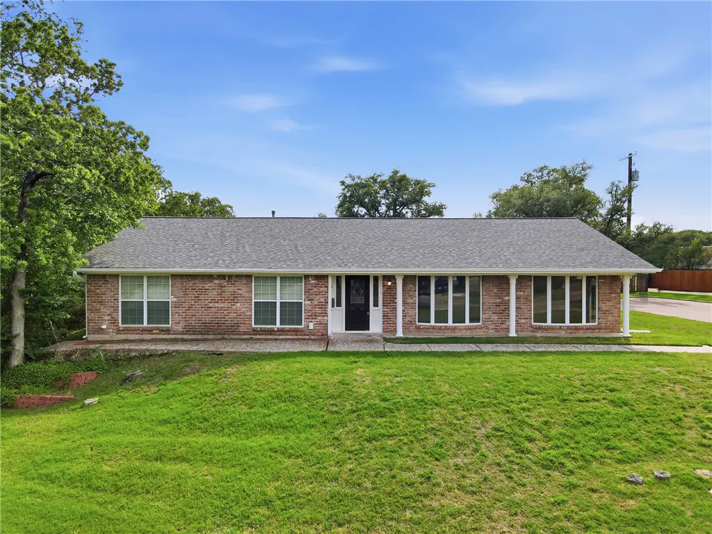 A single-story brick house with a gray roof, white trim, and a black front door sits on a green lawn under a blue sky.