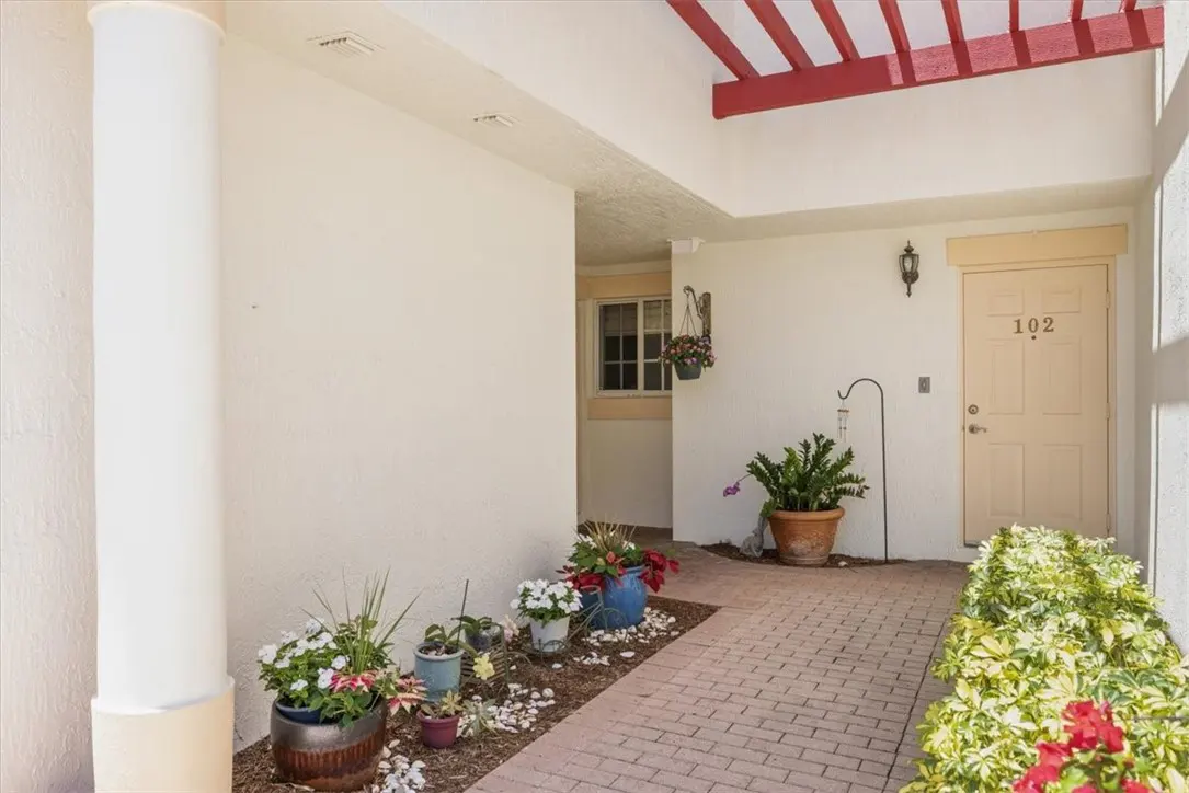 Exterior view of a condo entrance with potted plants, brick walkway, and a white column. Door number 102 is visible.