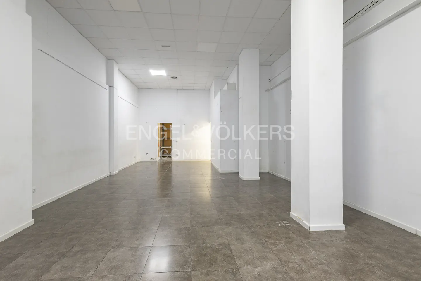 Empty commercial space with white walls, gray tile floor, and support columns. A doorway is visible in the back.