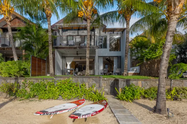 Two-story beach house with palm trees. Two red and white paddle boards on the sand. Stone path leads to the house.
