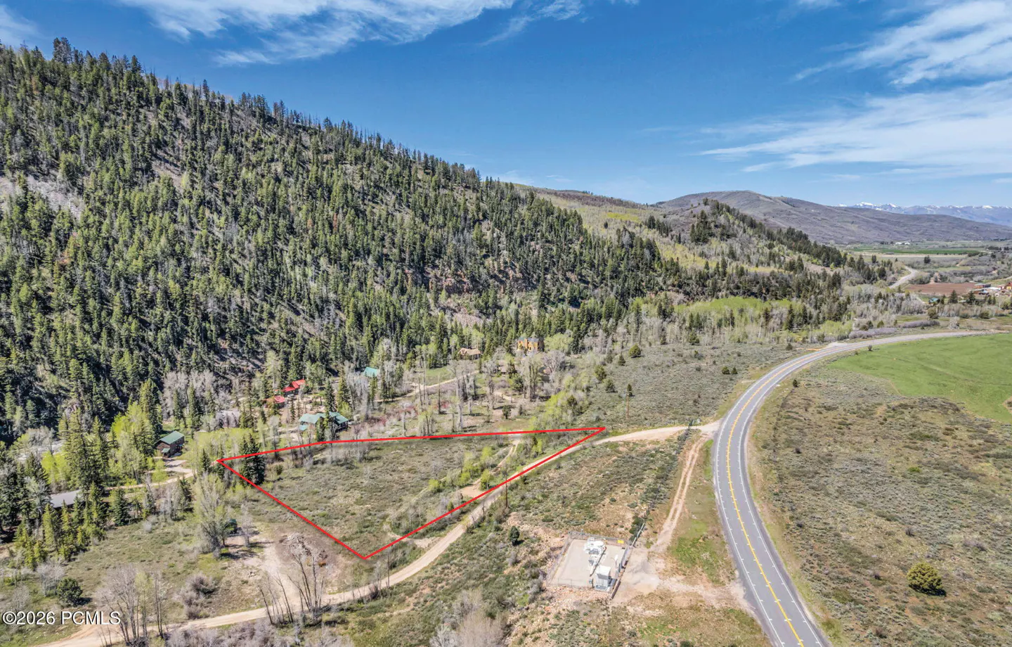 Aerial view of a vacant lot outlined in red, next to a road and a mountain covered in green trees under a blue sky.