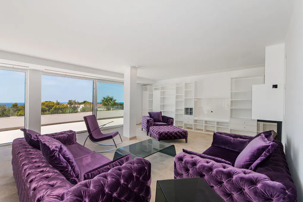 Bright living room with purple velvet sofas, chair, and ottoman. White walls, built-in shelves, and ocean view through large windows.