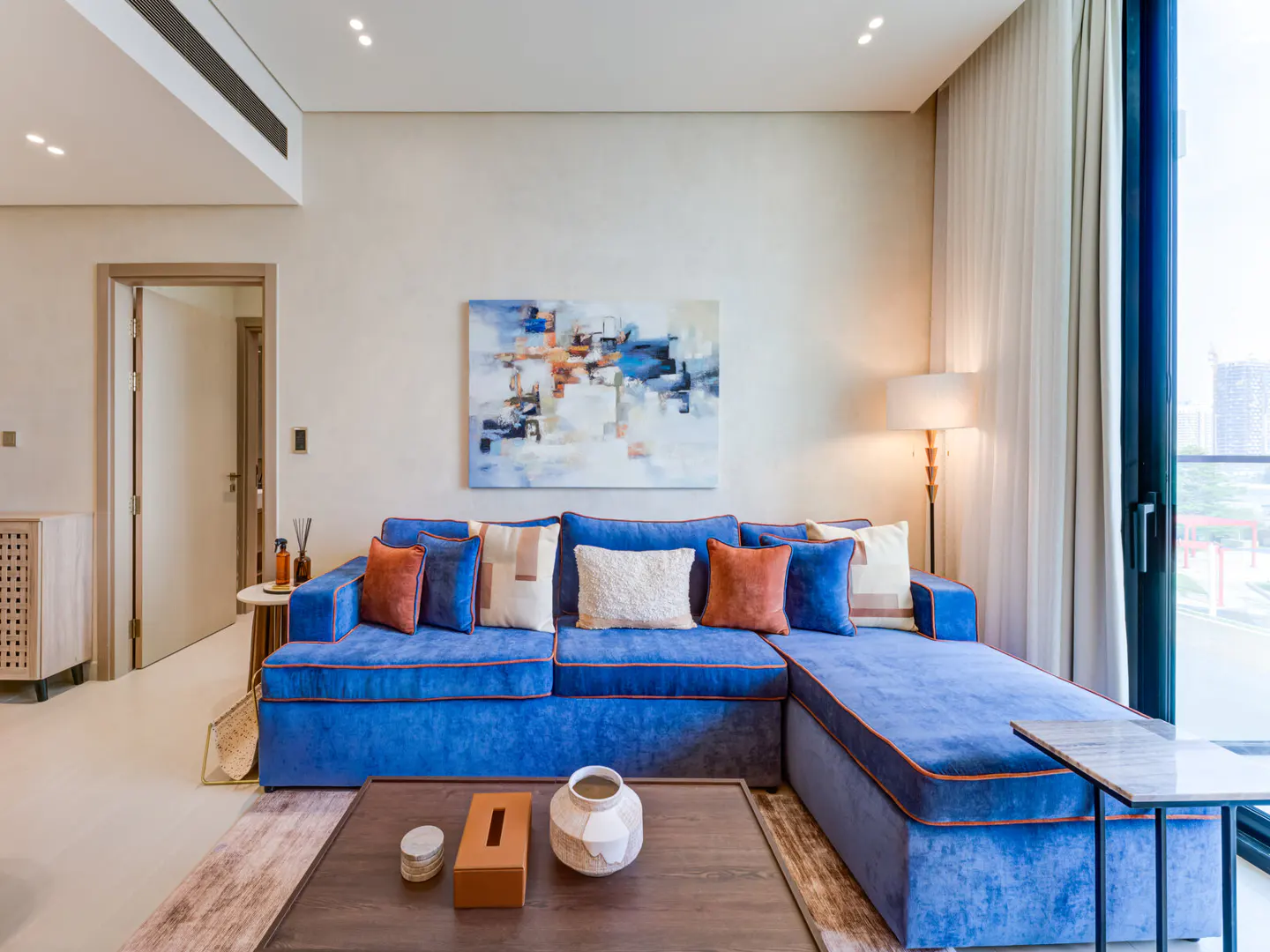 Living room with a blue sectional sofa, colorful pillows, and a dark wood coffee table with decorative items. An abstract painting hangs on the wall.