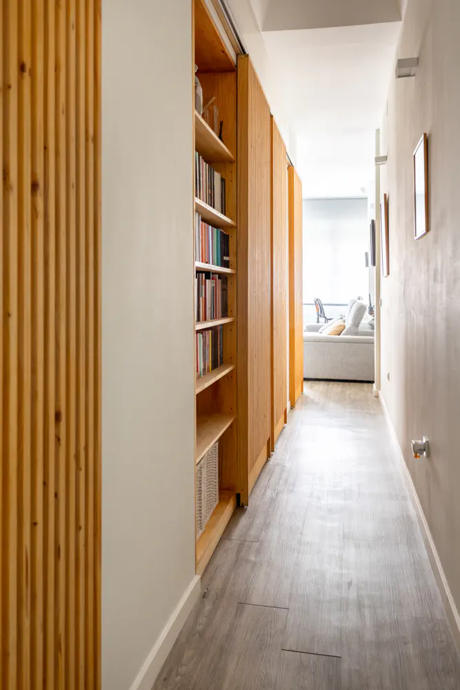 Hallway with light wood floors, a built-in bookcase with books, and a view into a living room with a white sofa.
