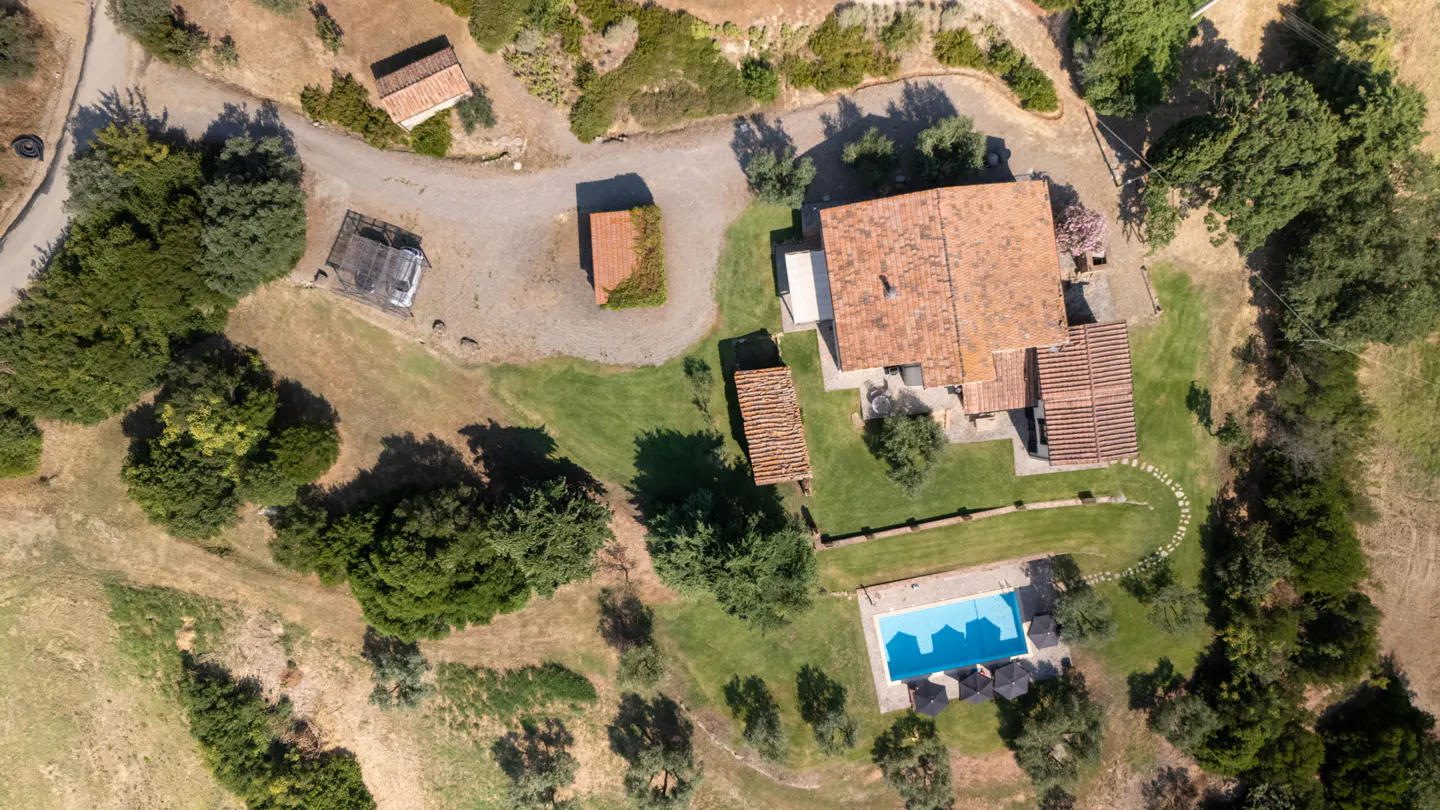 Aerial view of a large house with a red tile roof, a blue swimming pool, and green lawns surrounded by trees.