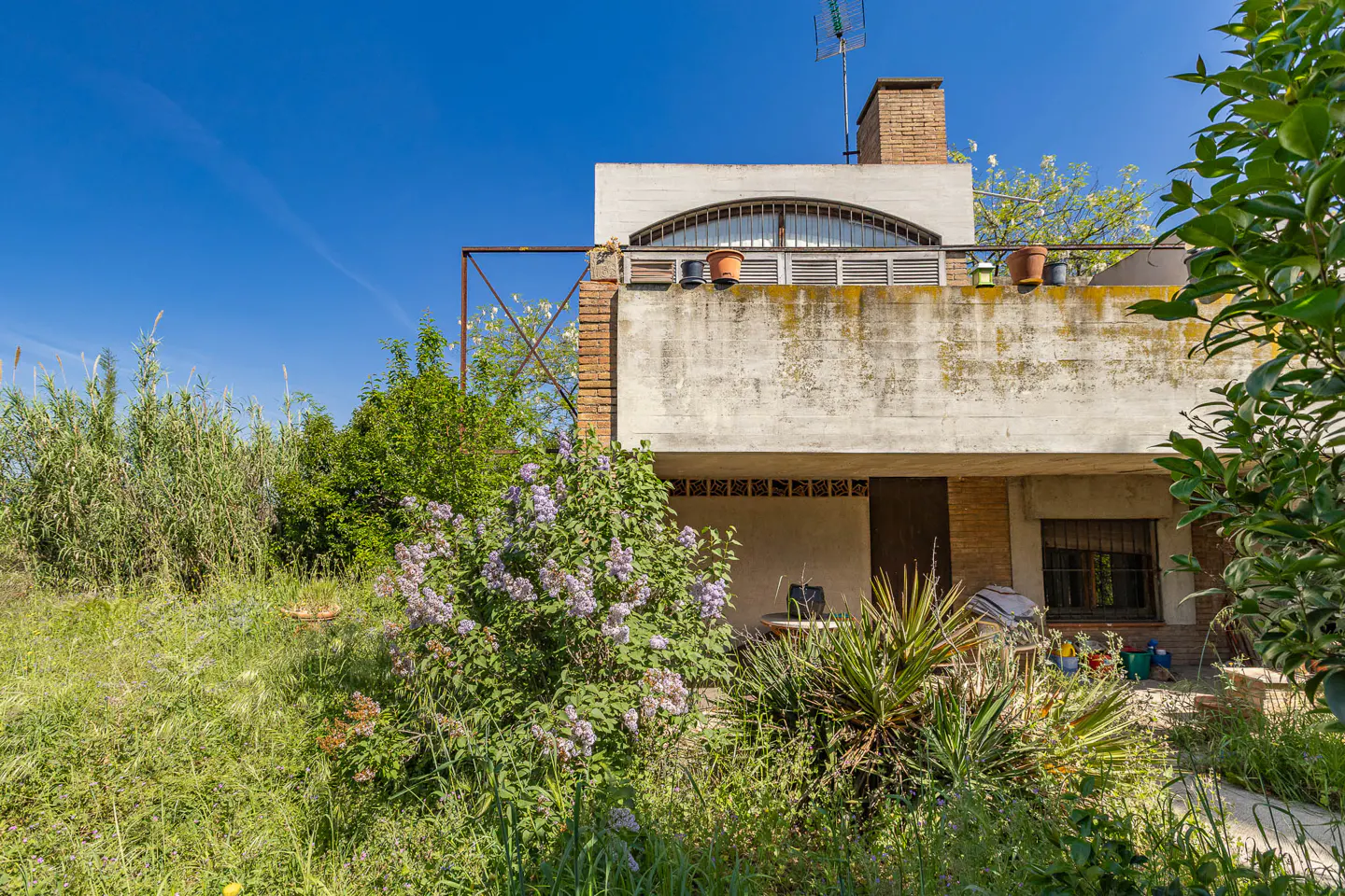 Exterior of a two-story house with a balcony, brick chimney, and overgrown garden under a blue sky.