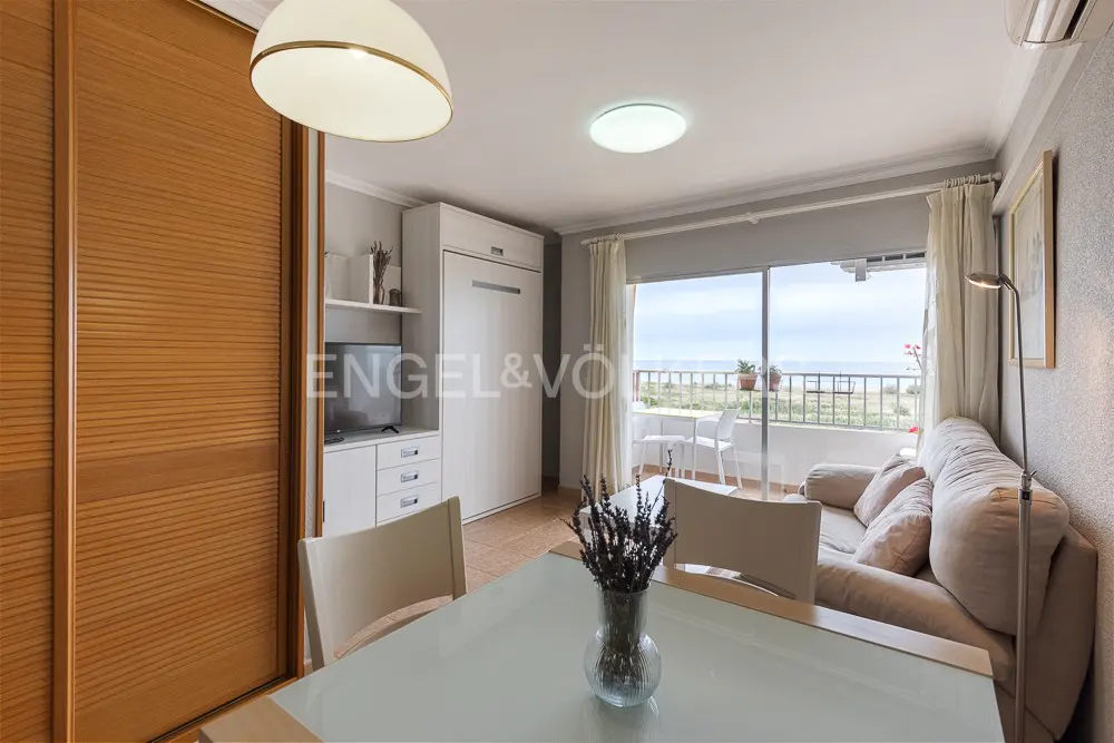 Modern living room with dining table, beige sofa, and glass sliding doors opening to a balcony with a sea view.