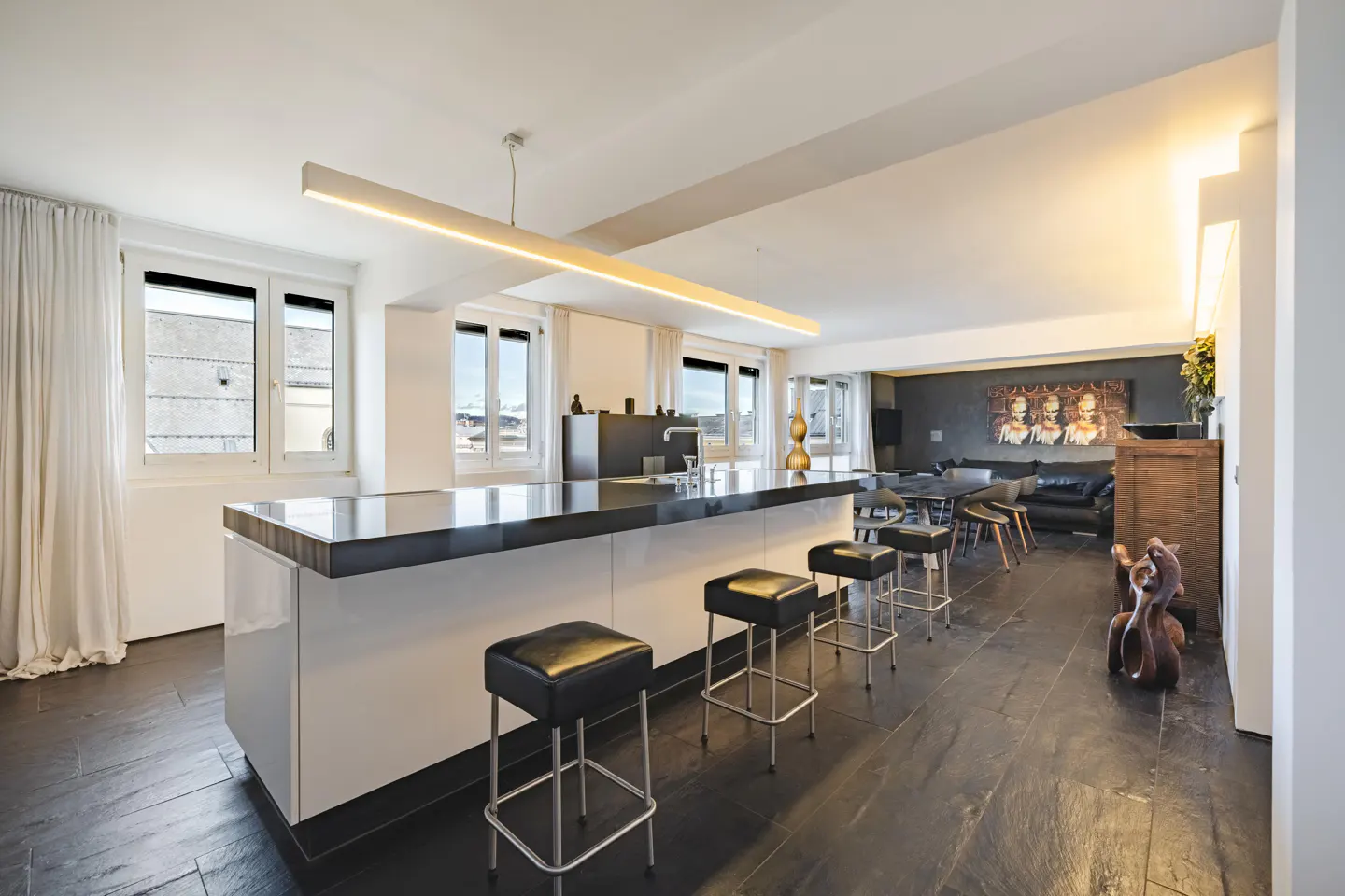 Bright, modern kitchen with a black countertop island and four black leather stools. Windows with white curtains and a living area in the background.
