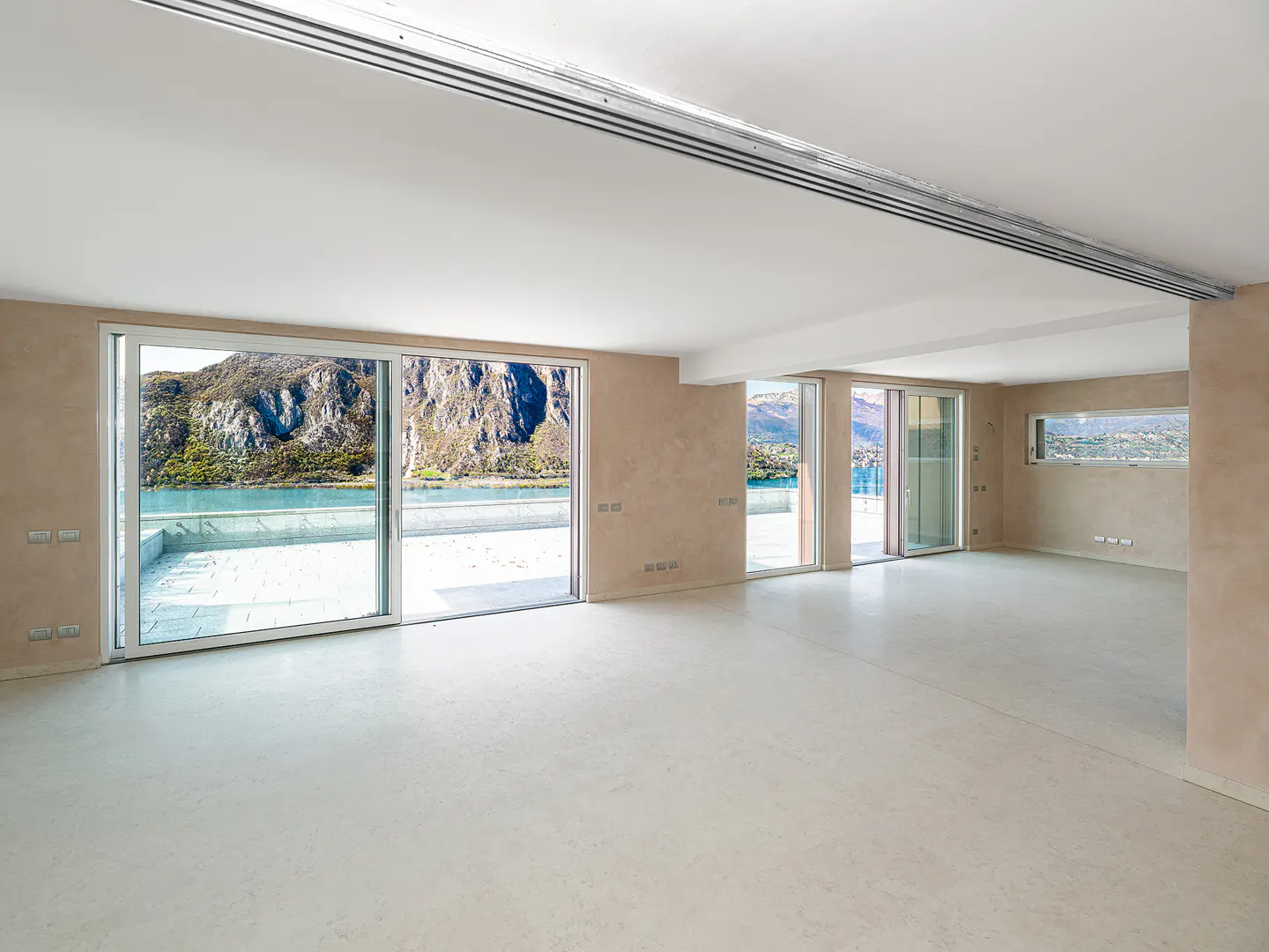 Bright, empty room with beige walls and white floor. Large sliding glass doors open to a balcony with a lake and mountain view.