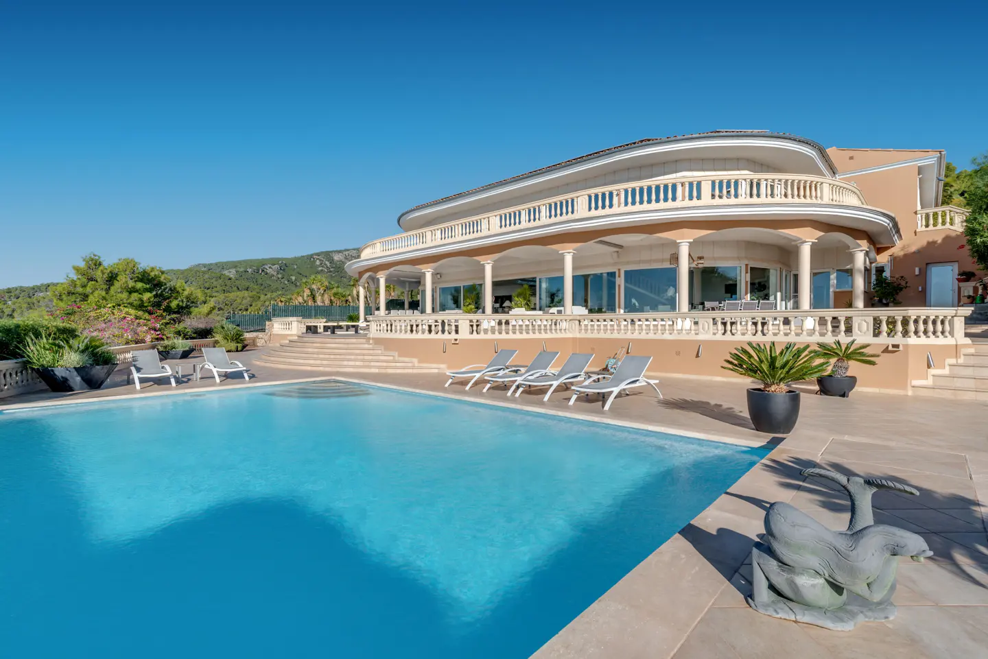 Luxury villa with a blue pool, lounge chairs, and a stone sculpture under a clear blue sky.