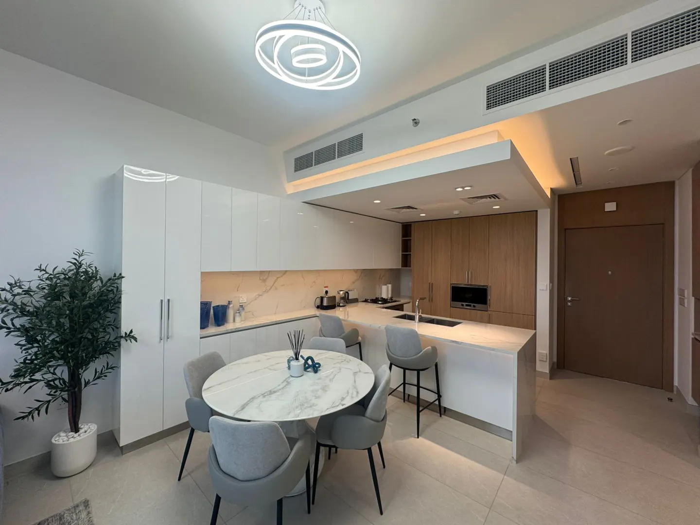A modern kitchen with white cabinets, marble countertops, and a round dining table with gray chairs. A decorative tree is on the left.