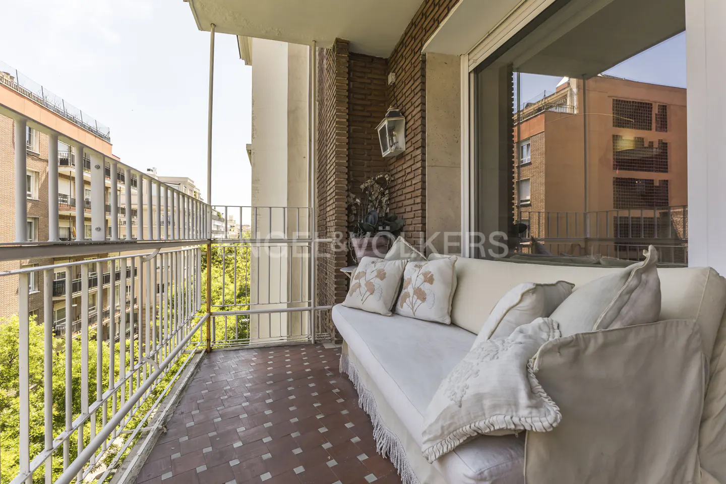 A cozy balcony with a white sofa, decorative pillows, and a view of city buildings. The floor is tiled in a red and white pattern.
