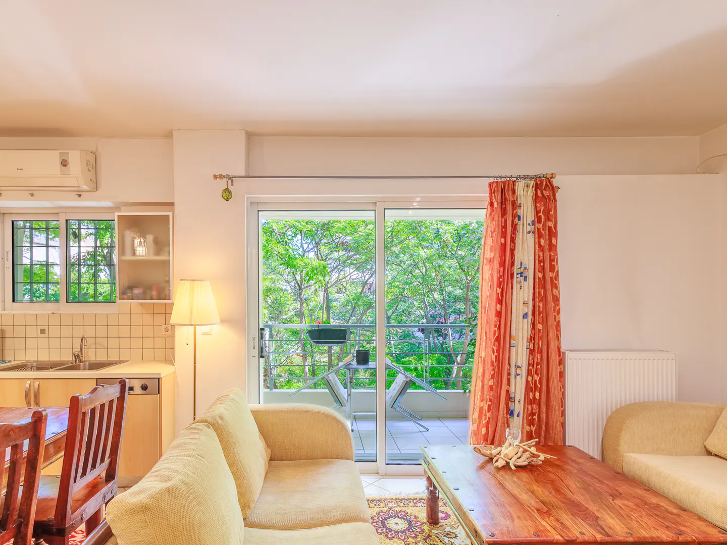 Bright living room with beige sofas, a wooden coffee table, and a balcony with green trees visible through sliding glass doors.
