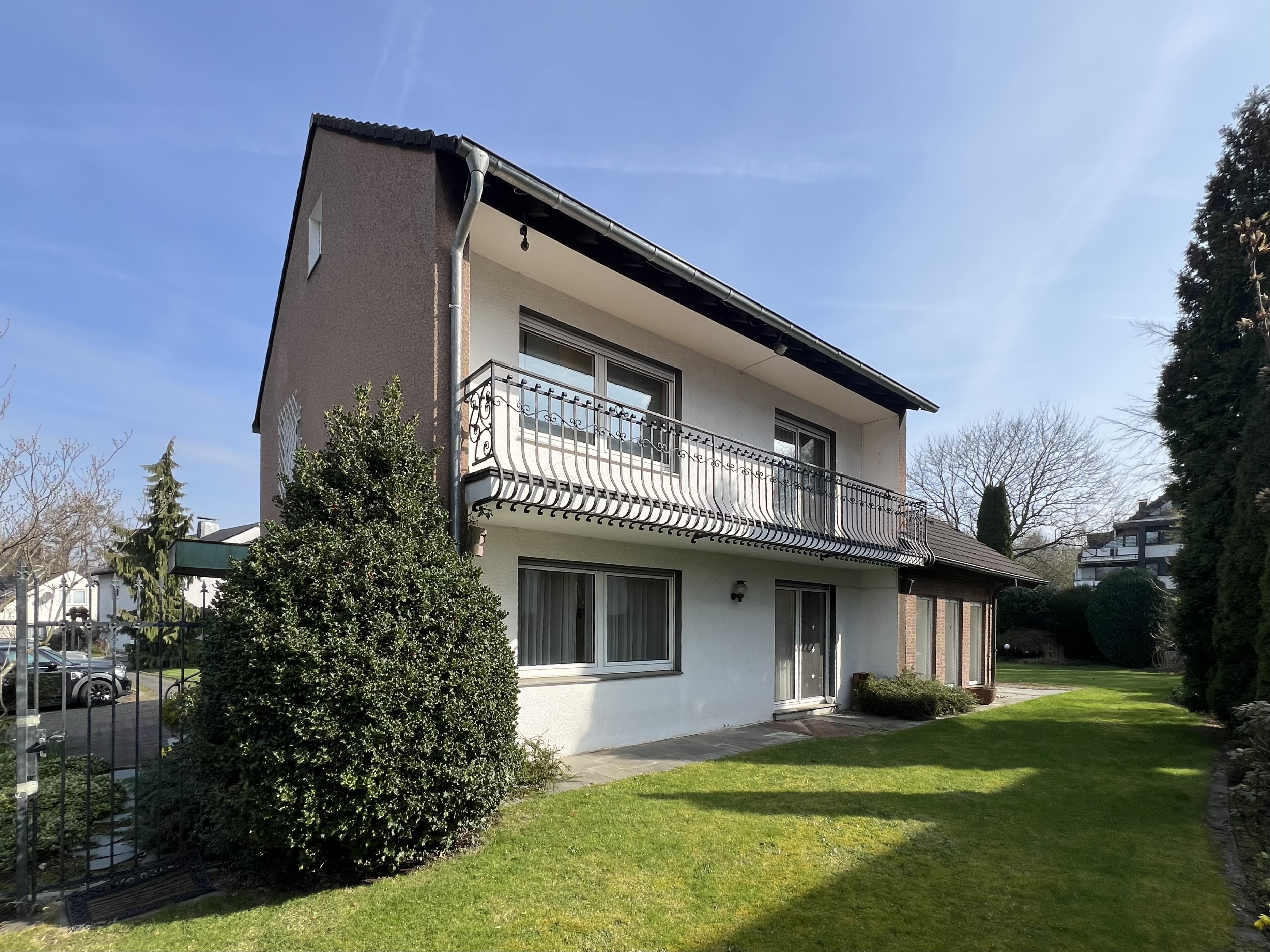 Two-story house with a brown and white exterior, black roof, and wrought iron balcony, surrounded by a green lawn and trees.