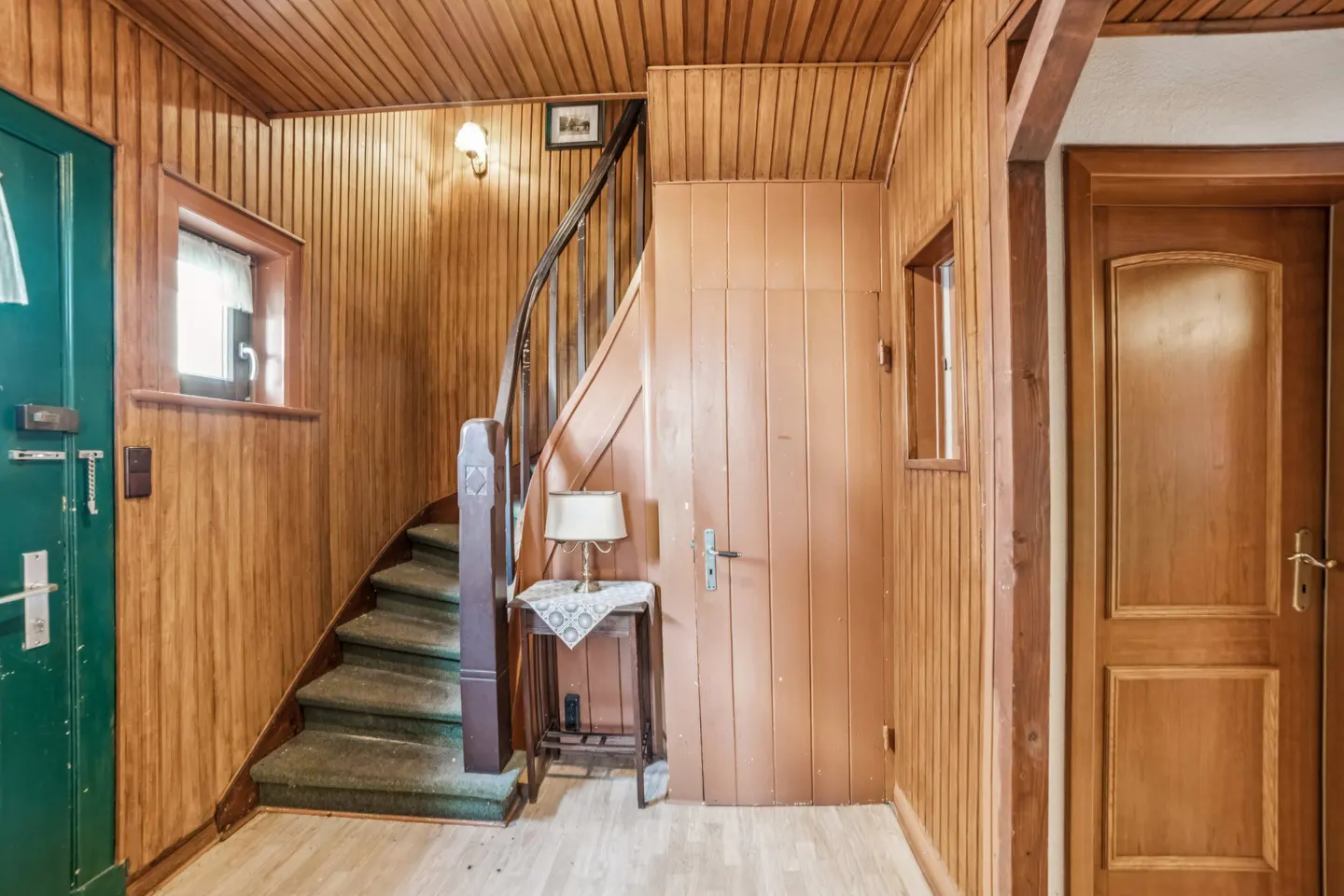 Interior view of a wood-paneled hallway with a staircase, green front door, and a small table with a lamp.