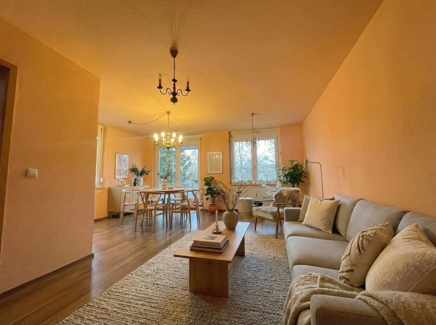 A wide shot of a peach-colored living and dining room with wood floors and a light gray sofa. A dining table and chairs sit near a window.