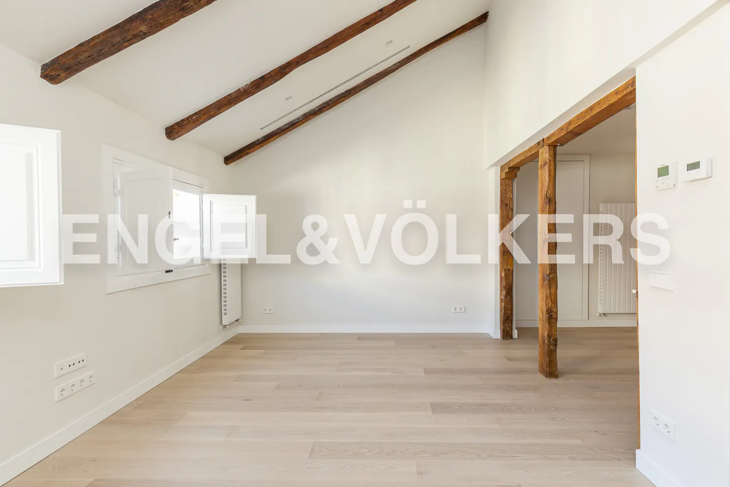 Bright, empty room with light wood floors, white walls, and exposed wood beams on the ceiling and as a doorway frame.