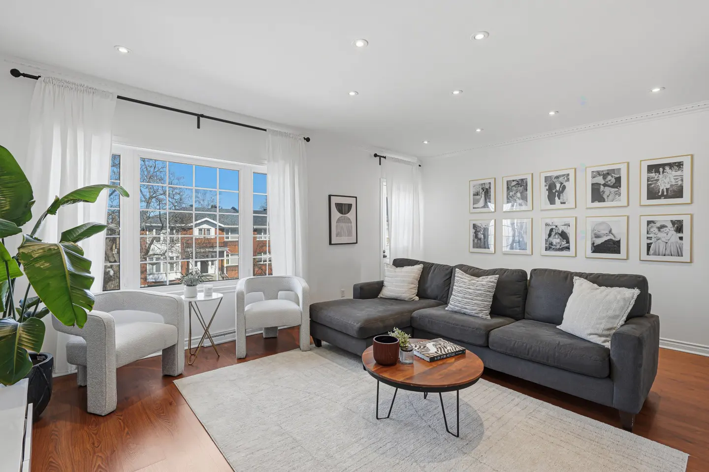 Bright living room with a gray sectional sofa, two white chairs, and a gallery wall of framed photos. A large window with white curtains lets in natural light.