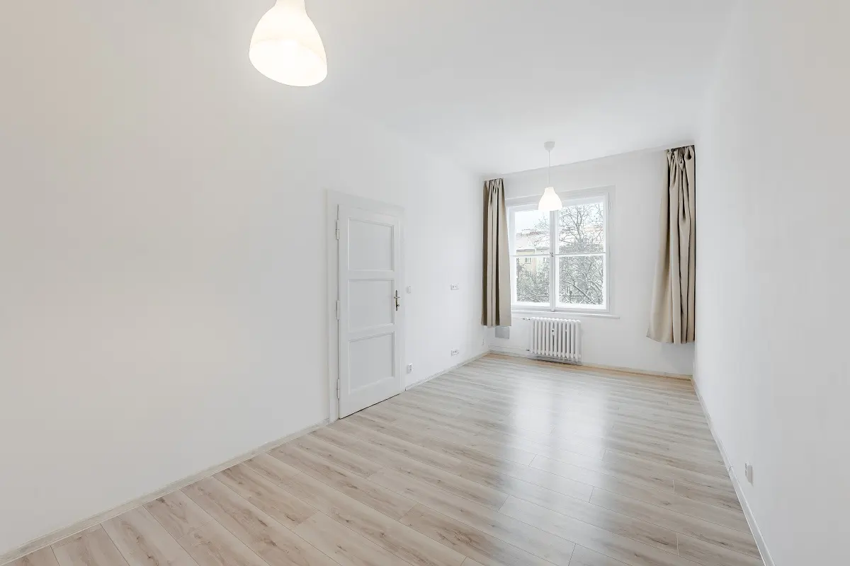 Bright, empty room with white walls, wood floor, and a window with beige curtains. A white door is on the left.