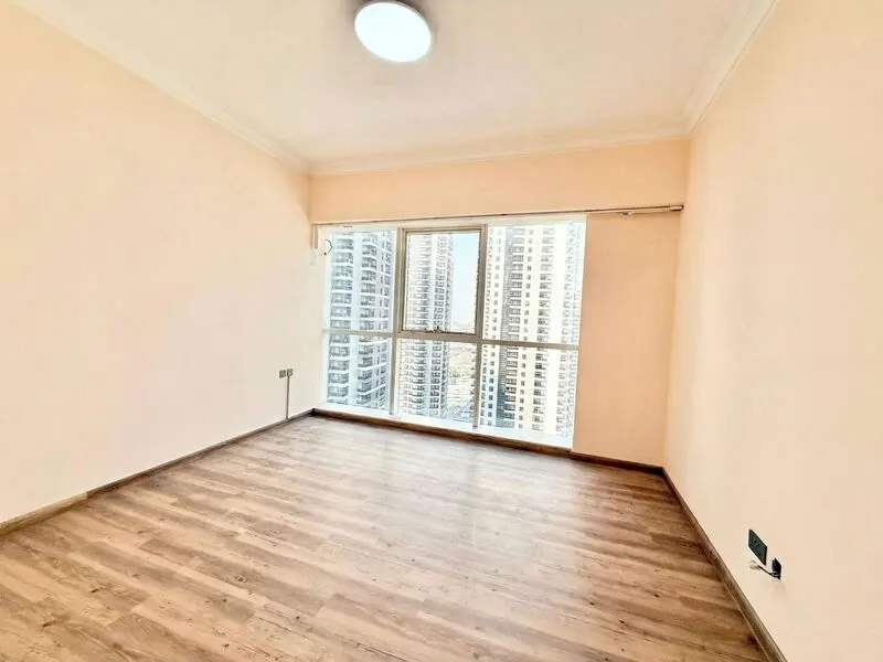 Empty room with light peach walls, wood-look flooring, a ceiling light, and a large window overlooking city skyscrapers.