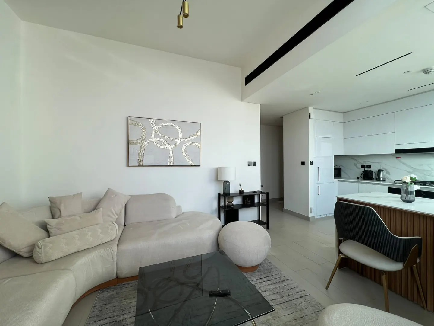 Modern open-plan living room with a beige curved sofa, glass coffee table, and a sleek white kitchen with a breakfast bar.