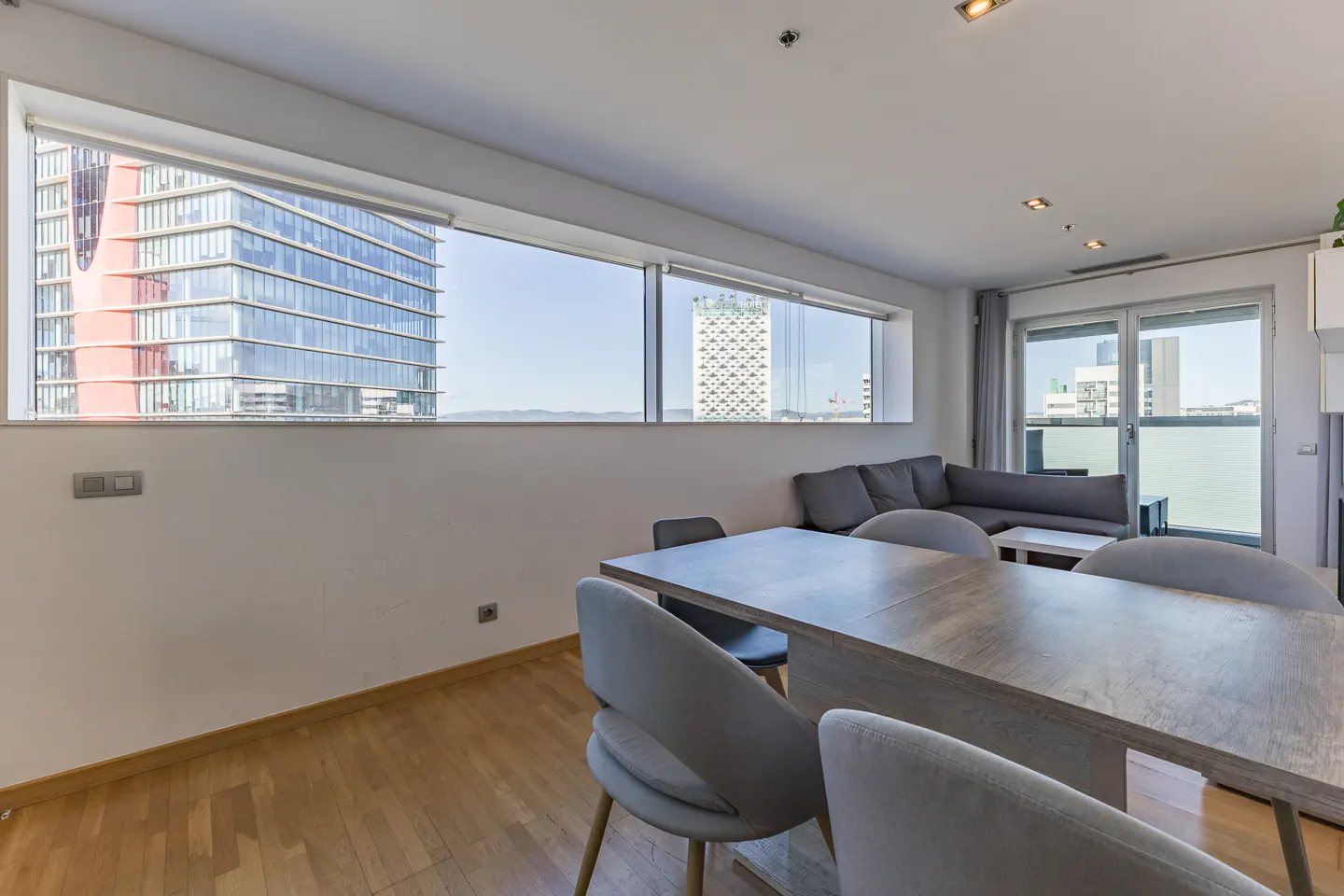 Bright apartment interior with a wood table, gray chairs, and a gray sofa. Large windows offer a city view.