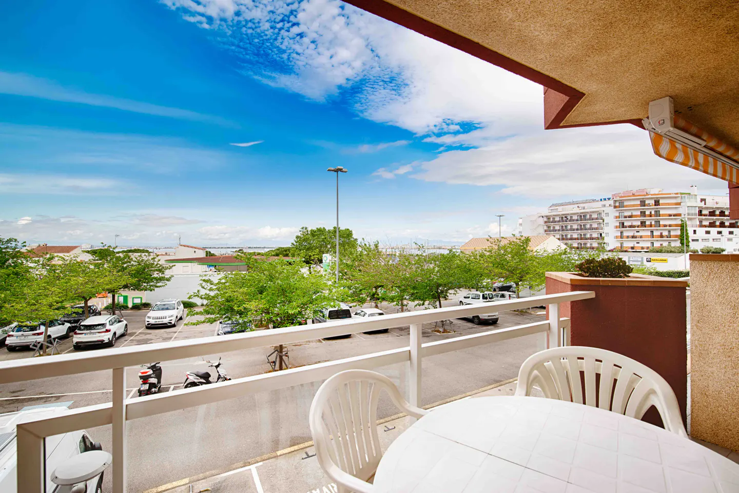 Balcony view with white table and chairs overlooking a parking lot, trees, and buildings under a blue sky.