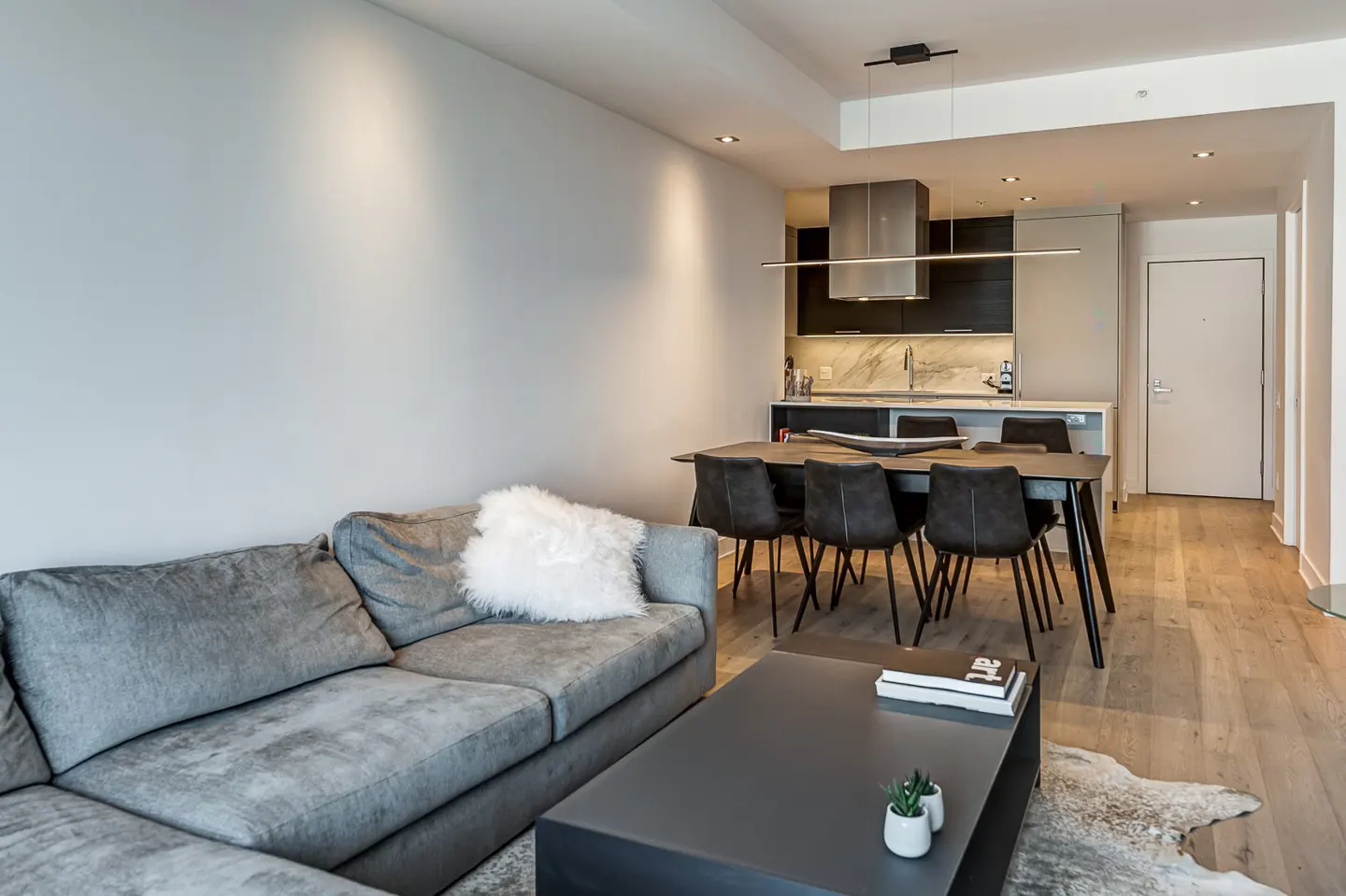 Modern open-concept living area with a grey sofa, coffee table, and dining set leading to a sleek kitchen with dark cabinets.