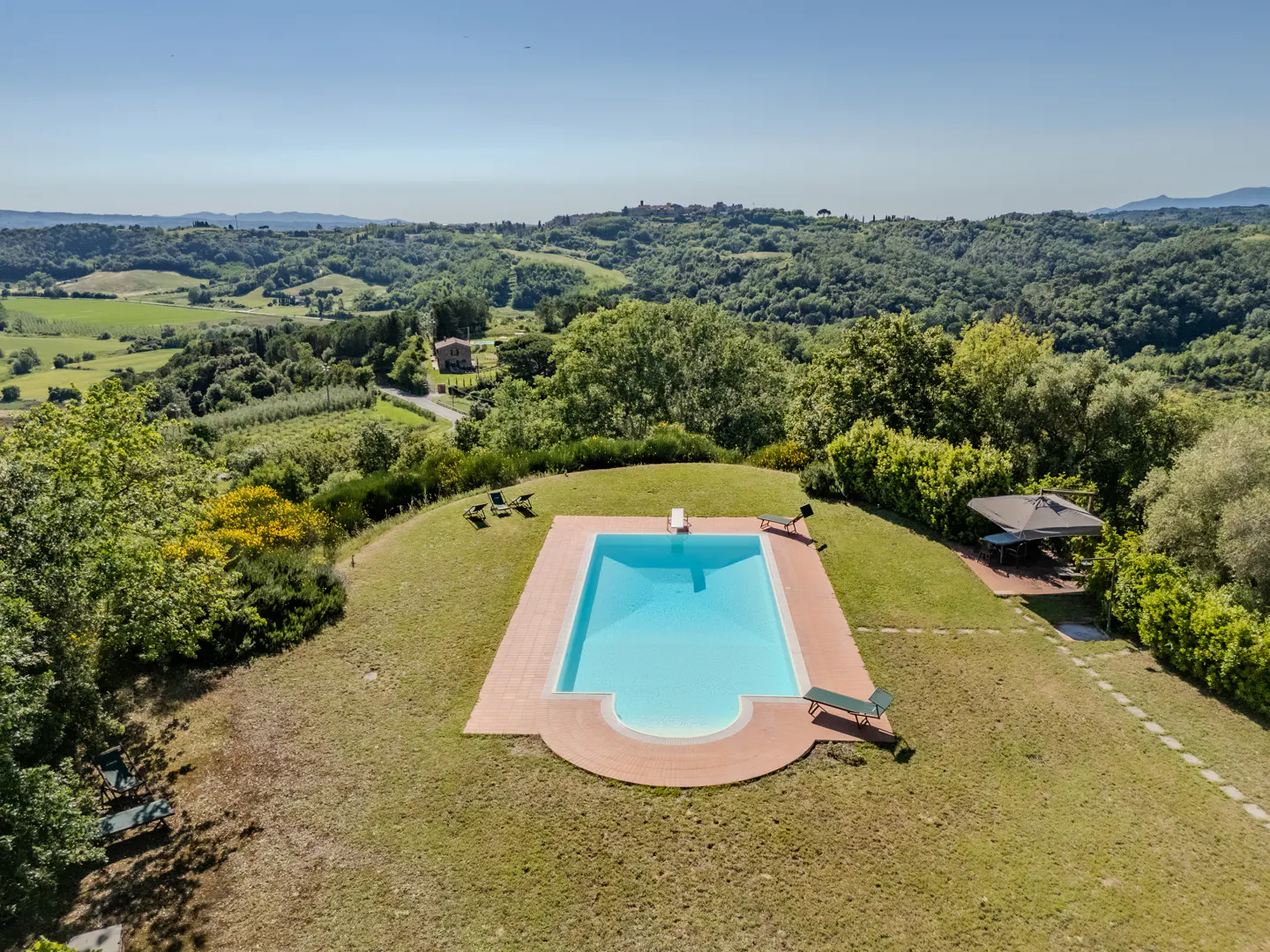 Aerial view of a rectangular pool with a diving board, surrounded by green grass, trees, and rolling hills in the background.