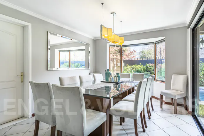 Bright dining room with a glass-topped table, white chairs, and a yellow chandelier. A large window overlooks a green garden.
