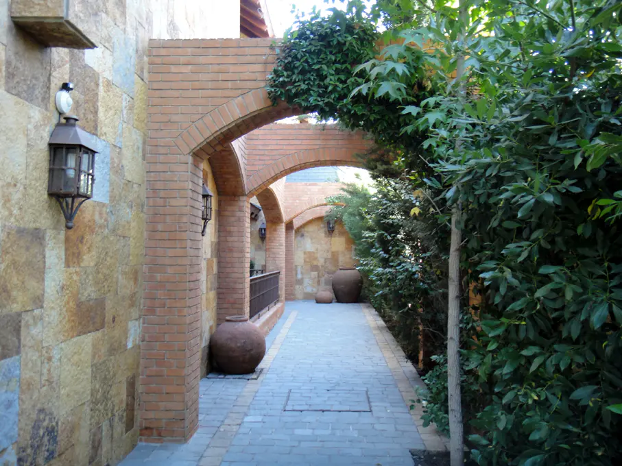 A brick archway leads to a stone-paved path lined with greenery and large clay pots. A stone wall with a lantern is on the left.