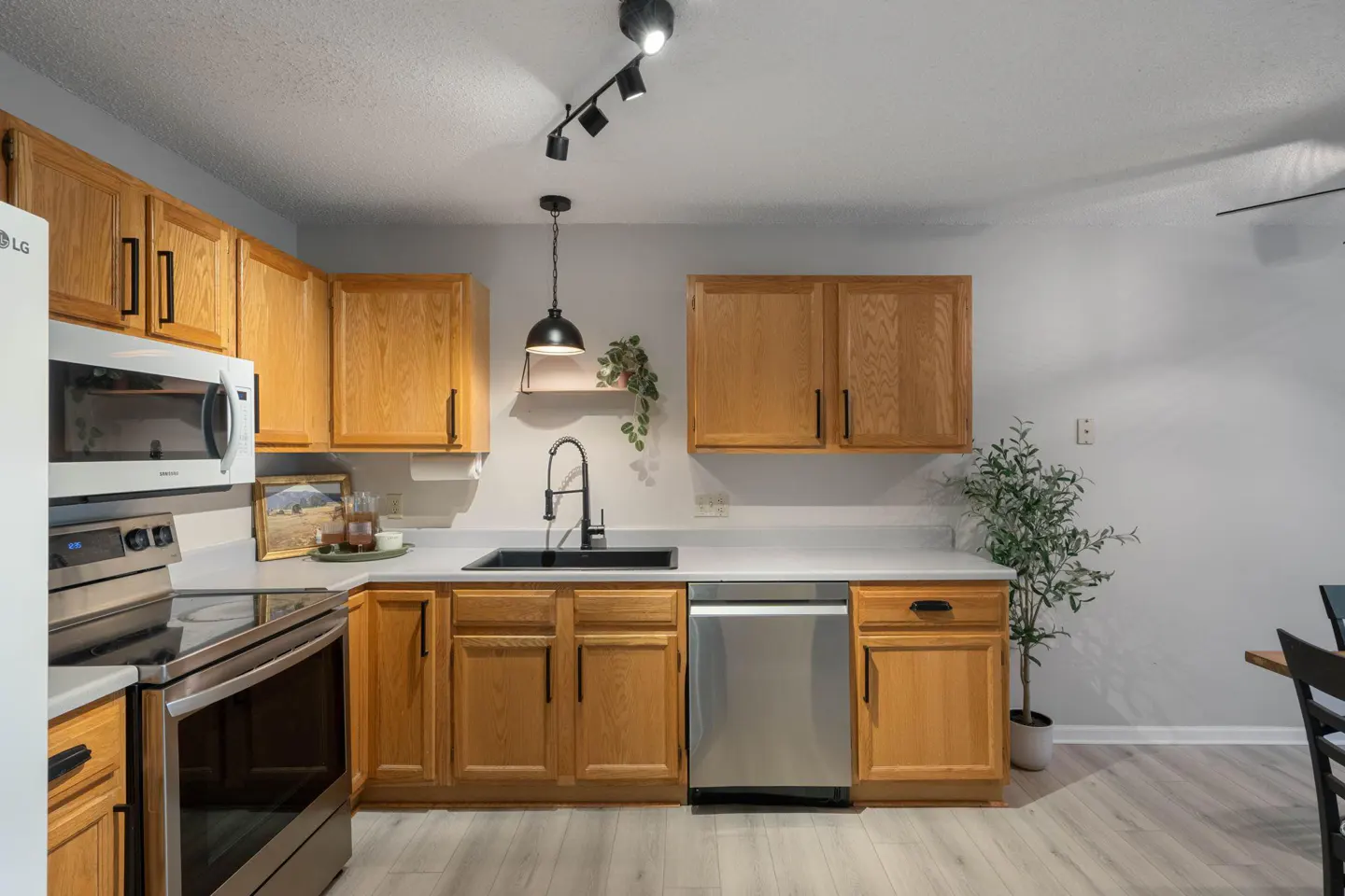 A bright kitchen with light wood cabinets, stainless steel appliances, and a black sink. Track lighting and a pendant lamp illuminate the space.