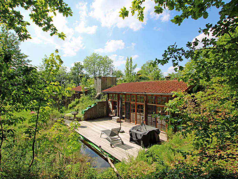 A rustic cabin with a red-tiled roof and wooden deck sits beside a small pond, surrounded by lush trees under a blue sky.