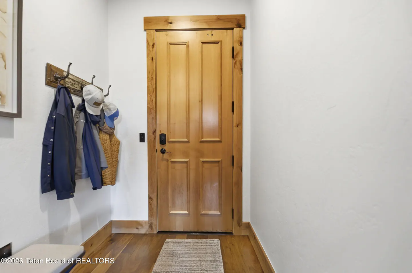 Entryway with a wooden door, hardwood floors, and a coat rack holding jackets and hats on a white wall.