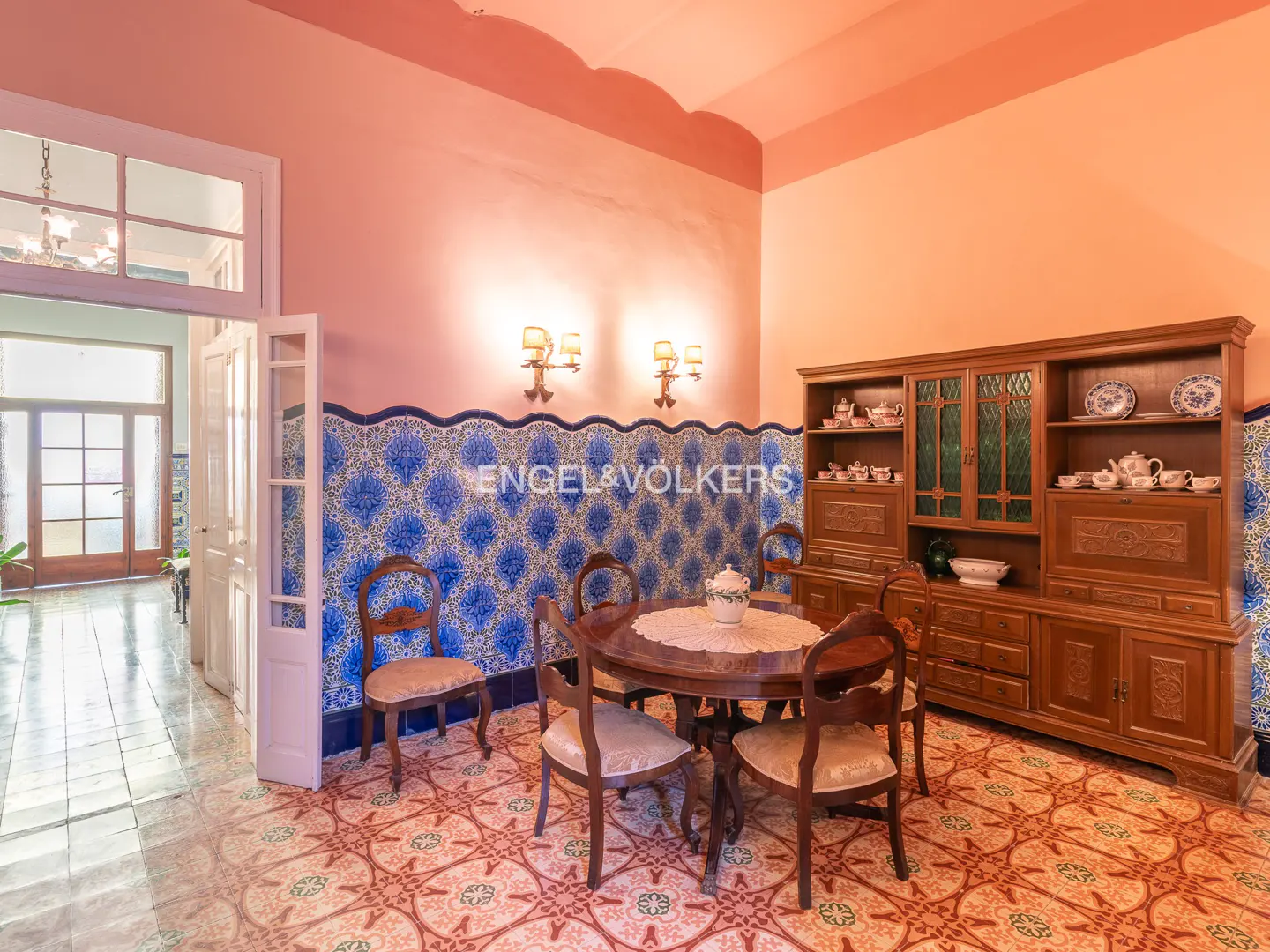 A dining room with a round wooden table, chairs, and a large wooden cabinet. Blue patterned tiles line the walls. The walls are pink.