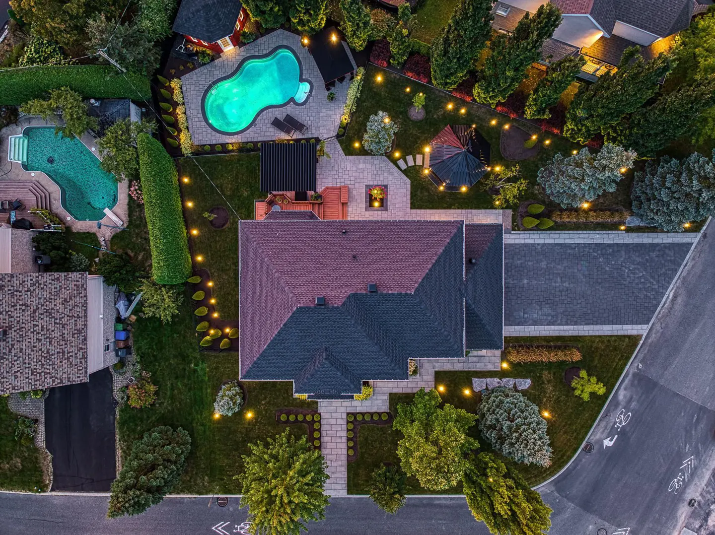 Aerial view of a house with a red and gray roof, a driveway, and a backyard with two pools and string lights.