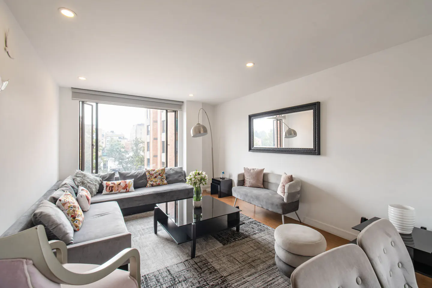 Bright living room with a gray sectional sofa, black coffee table, and large window overlooking a city view. A mirror hangs above a small sofa.