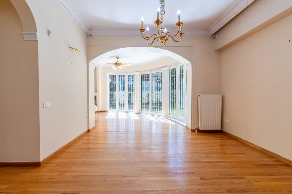 Empty room with hardwood floors, cream walls, and a gold chandelier. An arched doorway leads to a sunroom with white-framed windows.