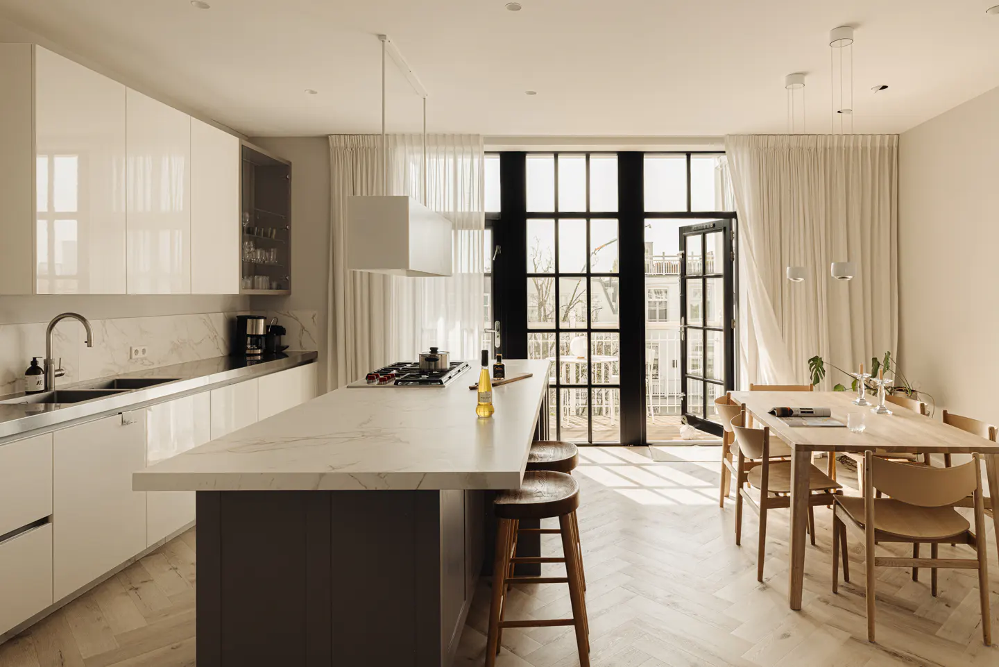 A bright, modern kitchen with white cabinets, a marble island, and a dining table near black-framed glass doors with white curtains.