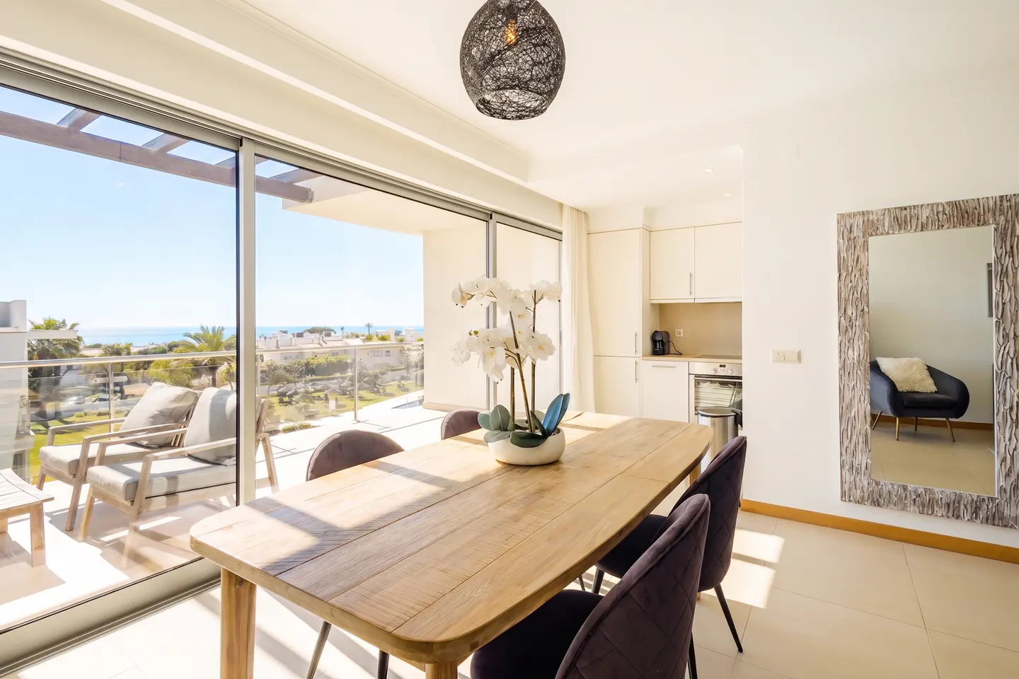 Bright dining area with a wooden table, purple chairs, and a view of the ocean from the balcony. A mirror reflects a chair.