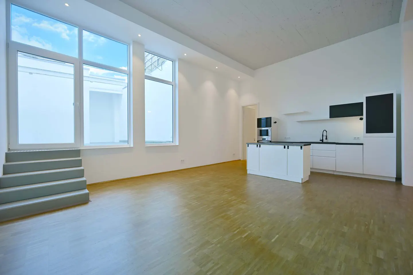 Bright, empty apartment with hardwood floors, white walls, and a modern white kitchen. Large windows with blue sky views and gray stairs.