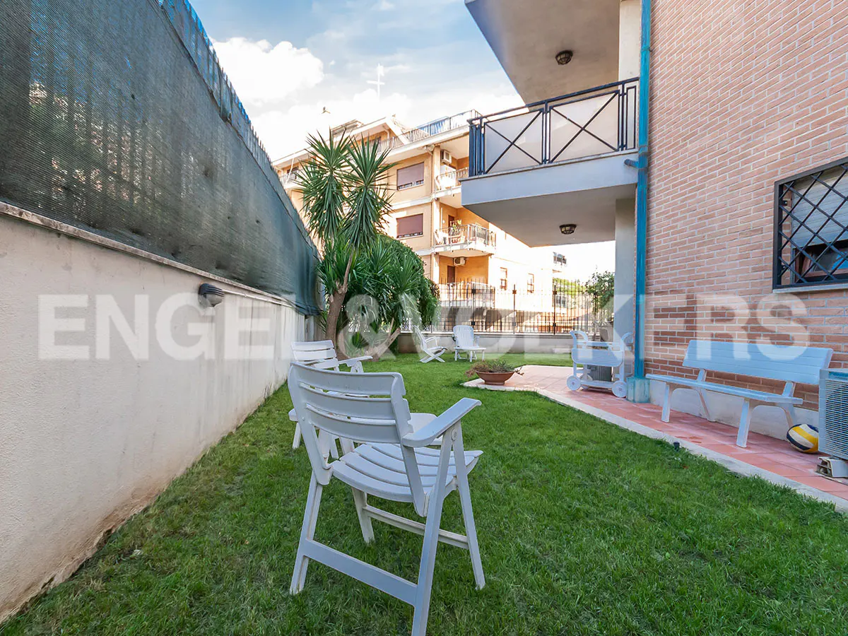 A backyard with green grass, white chairs, and a bench. A brick building is on the right.
