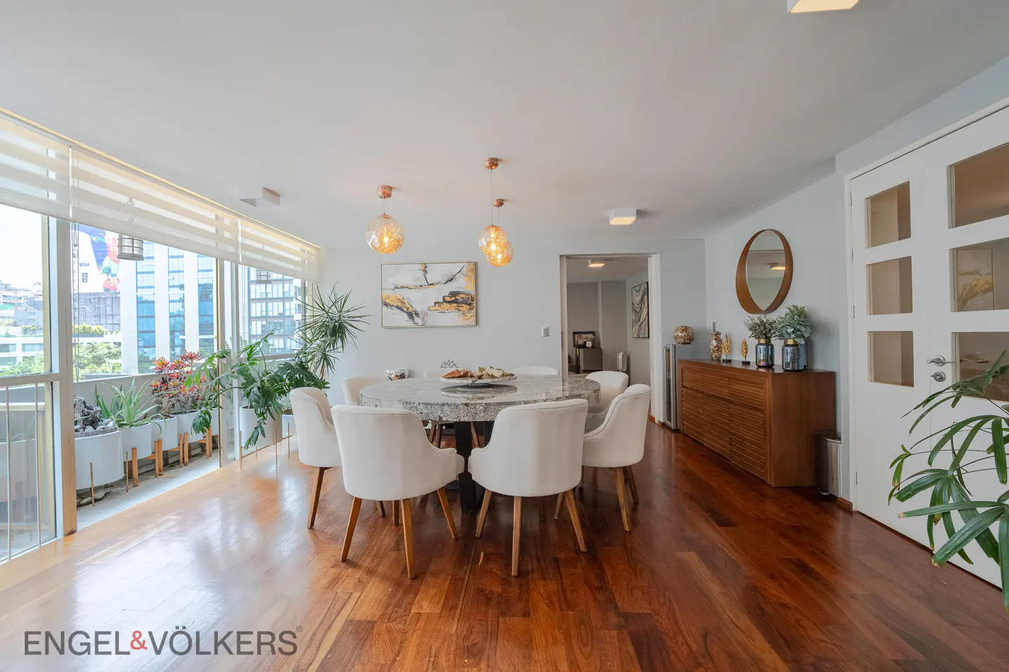 Dining room with a round stone table, white chairs, hardwood floors, and a balcony view through large windows.