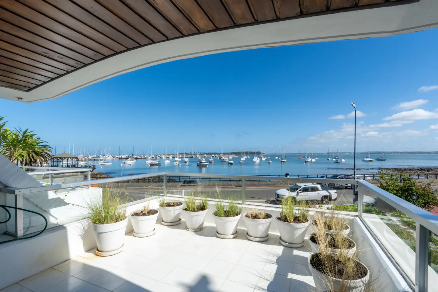 Balcony with white tiled floor, potted plants, and a glass railing overlooking a harbor filled with boats under a blue sky.