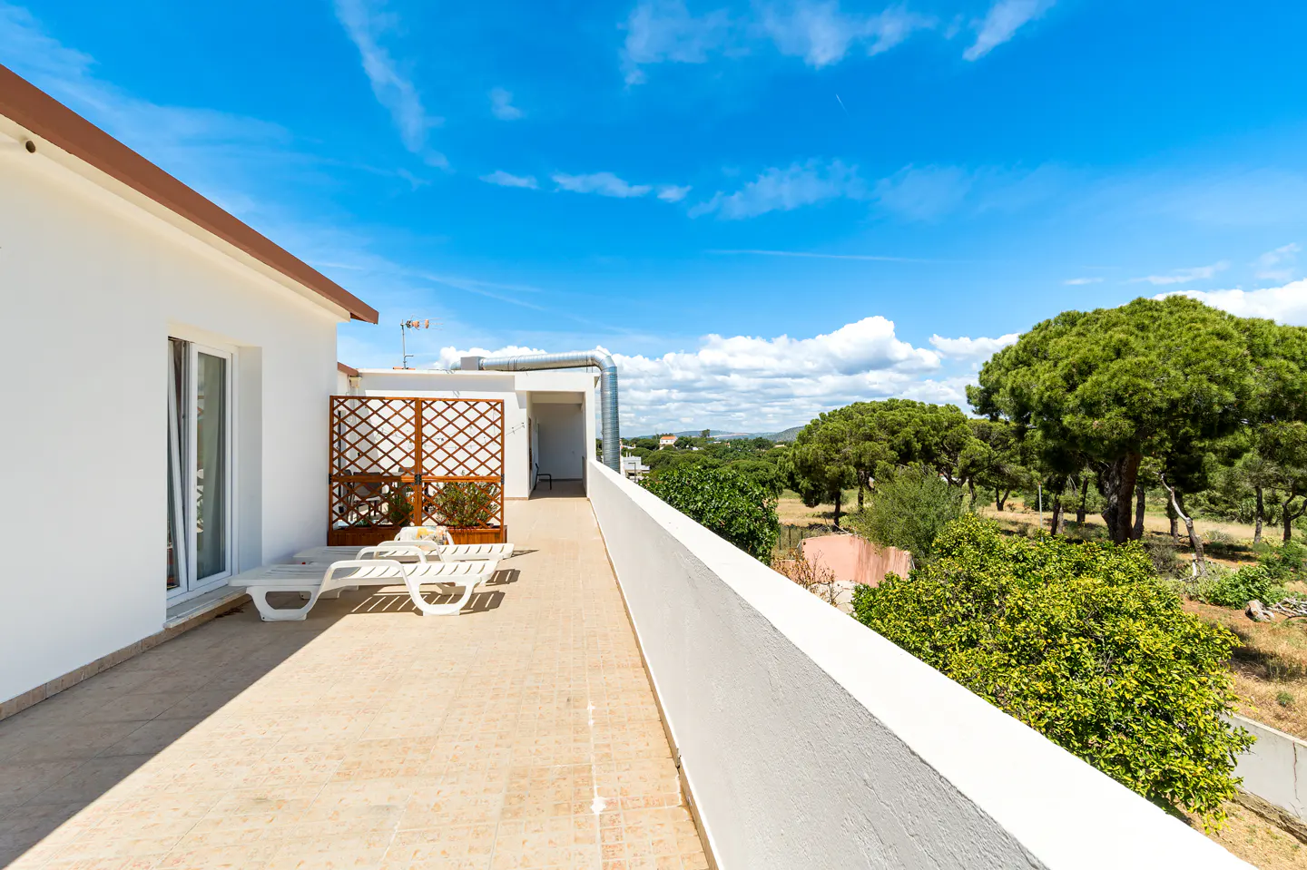 Balcony view with white lounge chairs, a wooden trellis, and a white building against a blue sky with trees in the background.