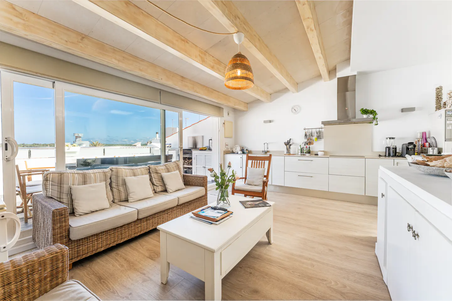 Bright open-plan living room with wicker sofa, white coffee table, and modern kitchen under vaulted wood-beamed ceilings.