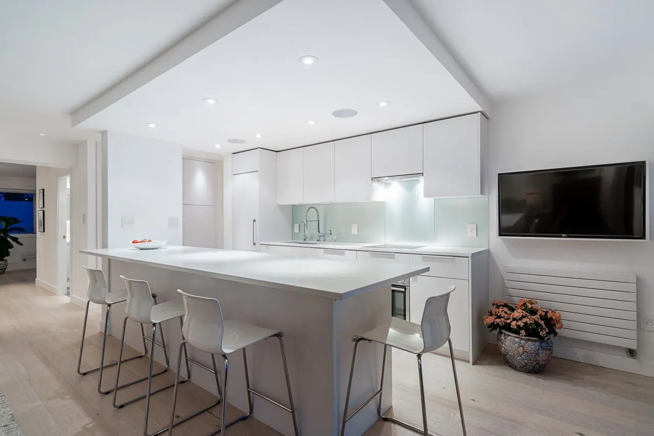 Bright, modern kitchen with white cabinets, island with stools, and a TV mounted on the wall. A flower pot sits below the TV.
