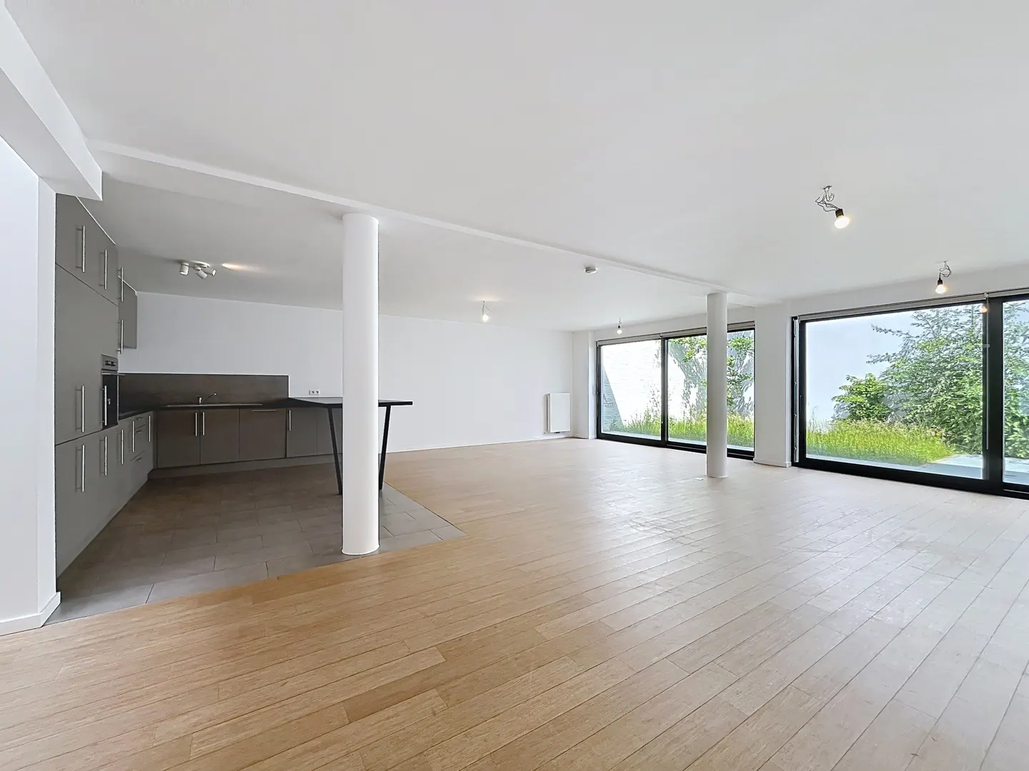 Modern open-plan living area with light wood floors, grey kitchen cabinets, white pillars, and large windows to a garden.
