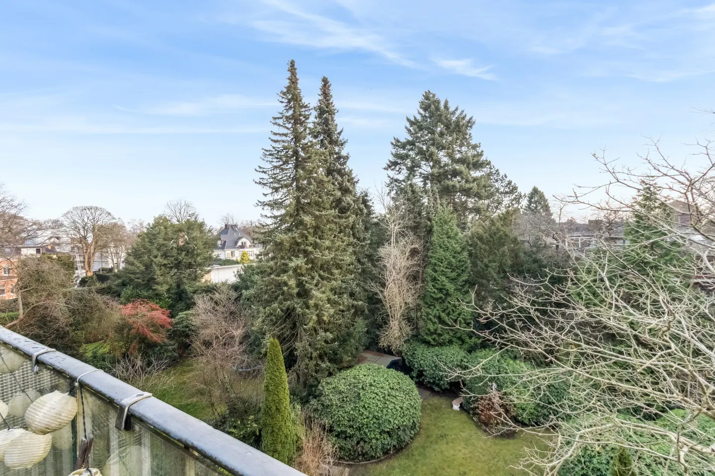 View from a balcony overlooking a lush green garden with tall trees under a blue sky.